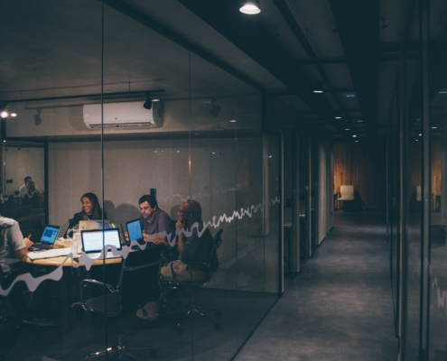 A dark image of a glass-walled office with people meeting with laptops at a table. Featured in an article on toxic cybersecurity culture.